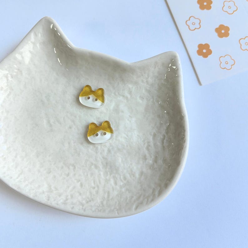 May include: Two small, cat-shaped buttons with a clear base and a gold-colored top section. Each button features two holes for attachment. They are resting inside a white, textured, cat-shaped ceramic dish next to a card with orange flower icons.