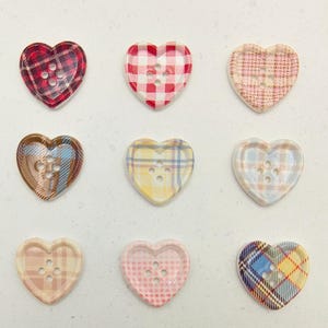 Op de afbeelding: Negen hartvormige knopen met ruitpatronen in verschillende kleuren. De knopen hebben een verscheidenheid aan kleuren, waaronder rood, roze, geel, blauw en bruin. Elke knoop heeft drie gaten om op te naaien.