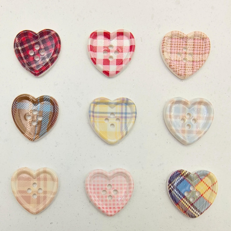 Op de afbeelding: Negen hartvormige knopen met ruitpatronen in verschillende kleuren. De knopen hebben een verscheidenheid aan kleuren, waaronder rood, roze, geel, blauw en bruin. Elke knoop heeft drie gaten om op te naaien.