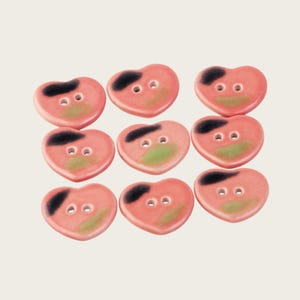 May include: A set of eight heart-shaped buttons in a smooth, glossy finish. Each button is primarily pink with a distinct black mark on the upper left side and a soft green patch on the lower centre. Every button features two central holes.