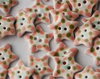 May include: A collection of ceramic star-shaped buttons with a white base and a pink and green speckled pattern. The buttons have two holes for sewing.