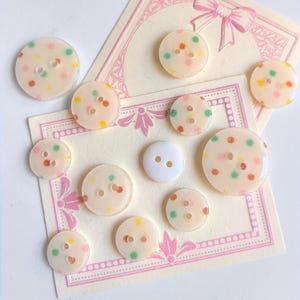 Peut inclure: Une collection de boutons décoratifs de différentes tailles et motifs. Les boutons sont principalement translucides avec des pois colorés. Un bouton est blanc uni. Les boutons sont disposés sur des cartes décoratives avec des accents roses.
