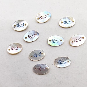 May include: Oval mother-of-pearl beads featuring a dachshund dog design. The dog illustrations are in red, blue, and black. Each bead has two holes for stringing. The beads have a pearlescent sheen.