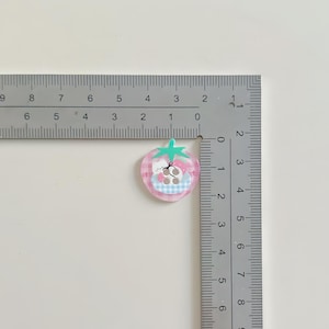 May include: A pink strawberry-shaped charm with a cartoon bear design. The charm has a green stem and a blue and white checkered pattern. The charm is approximately 1 inch in diameter.