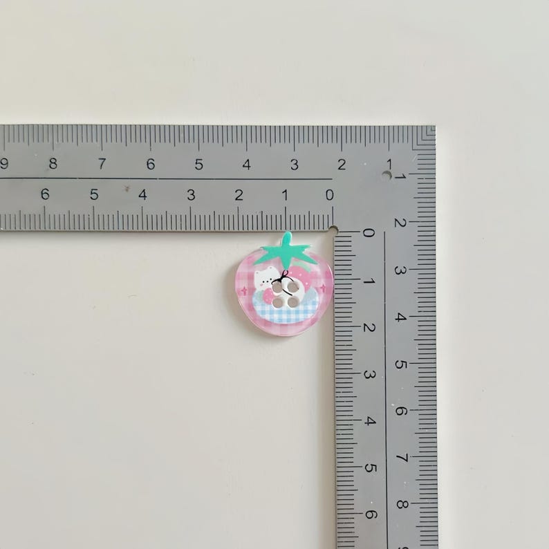 May include: A pink strawberry-shaped charm with a cartoon bear design. The charm has a green stem and a blue and white checkered pattern. The charm is approximately 1 inch in diameter.