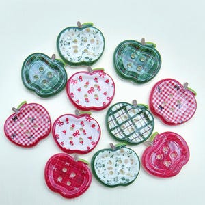 Può includere: Una collezione di bottoni decorativi a forma di mela in vari colori e motivi. I bottoni presentano disegni come stampe floreali, quadri e fiocchi. Ogni bottone ha un gambo verde ed è realizzato in materiale lucido.