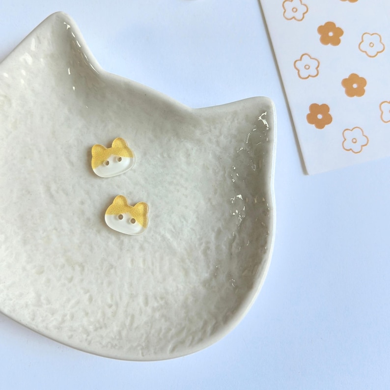May include: Two small, translucent cat-shaped buttons with a yellow top and white bottom, each featuring two holes for sewing. These buttons are resting on a textured, off-white ceramic dish shaped like a cat head, next to a white card with orange flowers.