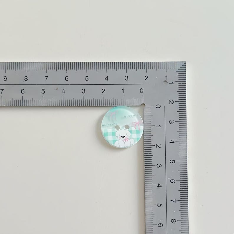 May include: A round, two-hole button featuring a light green and white checkered pattern with a white puppy illustration and a pink bow. A metal L-shaped ruler is positioned against the button, showing a diameter of approximately 0.75 inches.