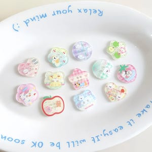 May include: A collection of colorful, transparent acrylic charms in various shapes, including hearts, flowers, and circles. Each charm features a cute bear or other character design. The charms are arranged on a white plate with blue text.