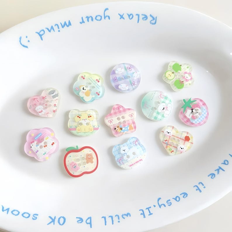 May include: A collection of colorful, transparent acrylic charms in various shapes, including hearts, flowers, and circles. Each charm features a cute bear or other character design. The charms are arranged on a white plate with blue text.