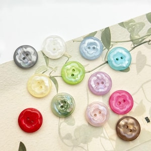 Peut inclure: Une collection de boutons irisés en forme de fleur, de différentes couleurs : gris, blanc, bleu, vert, jaune, violet, rose, rouge et marron. Chaque bouton comporte deux trous pour la couture. Les boutons sont disposés sur une surface claire.