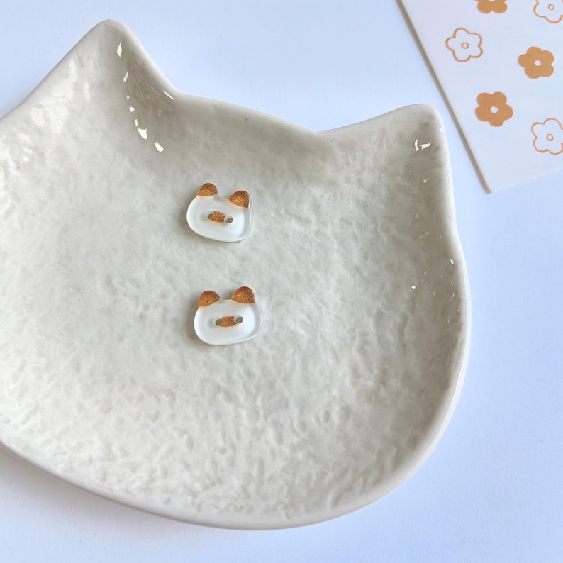 May include: Two small, translucent white bear-shaped charms with brown ears and facial details, resting on a textured, off-white ceramic dish shaped like a cat head. The dish has a rough, uneven surface and pointed ears at the top edge.