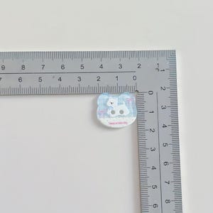 May include: A small, light blue and white cat-shaped button with a cartoon cat design. The button is approximately 1 inch wide. The cat is wearing a blue scarf and the button has the text "have a nice day".