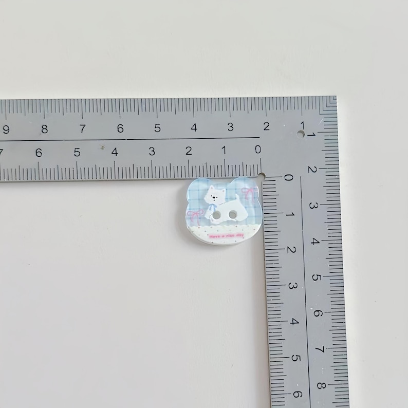 May include: A small, light blue and white cat-shaped button with a cartoon cat design. The button is approximately 1 inch wide. The cat is wearing a blue scarf and the button has the text "have a nice day".