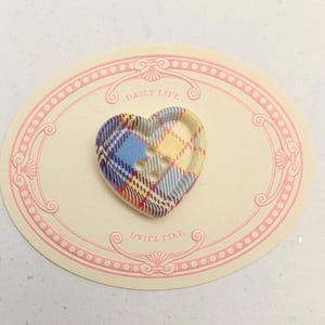 Op de afbeelding: Een hartvormige knoop met een blauw, geel en rood geruit patroon. De knoop heeft drie gaten en ligt op een cr&egrave;mekleurige ovale kaart met de woorden "DAILY LIFE" erop.