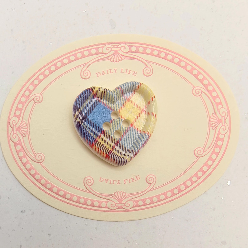Op de afbeelding: Een hartvormige knoop met een blauw, geel en rood geruit patroon. De knoop heeft drie gaten en ligt op een cr&egrave;mekleurige ovale kaart met de woorden "DAILY LIFE" erop.
