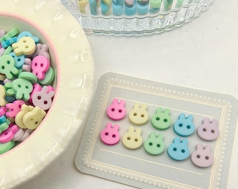 Pode incluir: Uma coleção de botões em forma de coelho em tons pastel, rosa, amarelo, verde, azul e roxo. Alguns botões estão numa tigela branca, enquanto outros estão dispostos num cartão decorativo. Um recipiente de vidro transparente com contas coloridas também é visível.