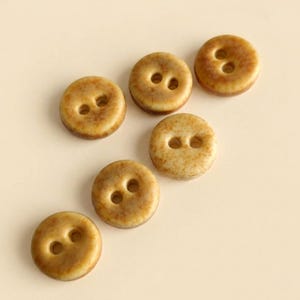 May include: Six round buttons with a smooth, glossy finish and a mottled tan and brown speckled pattern. Each button features two centered holes for attachment, displaying a consistent circular shape and a warm, earthy color palette throughout.