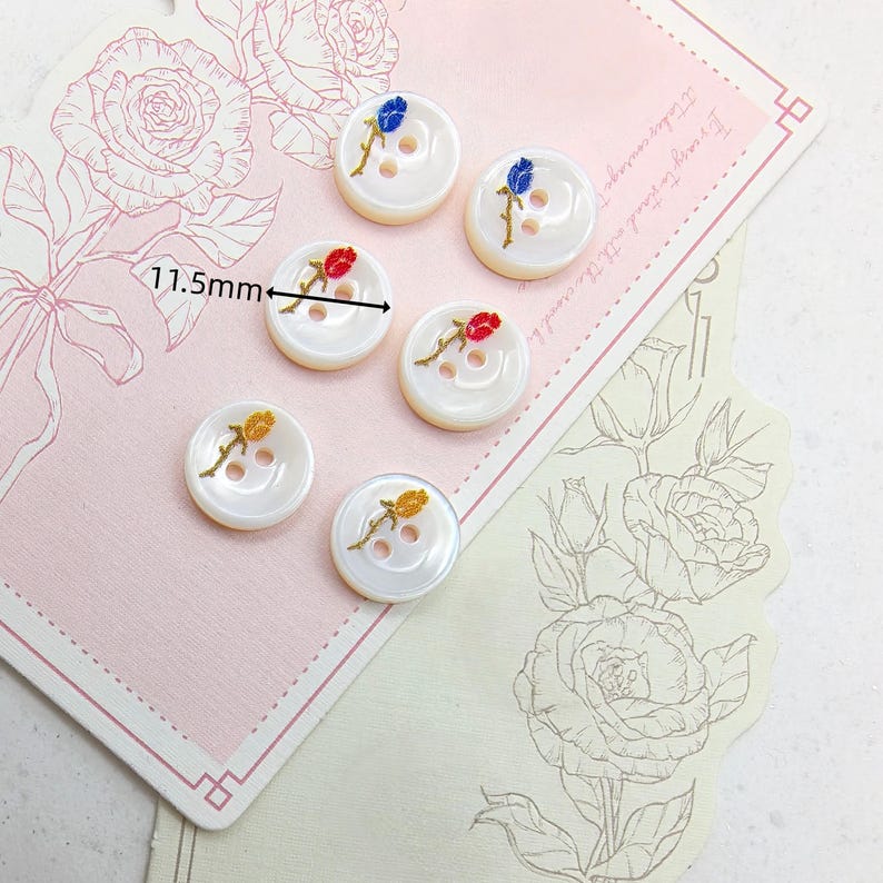 May include: Six white, iridescent buttons with embroidered rose designs in red, yellow, and blue. Each button measures 11.5mm in diameter. The buttons are arranged on a pink and cream floral-themed background.