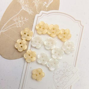 May include: A collection of eleven flower-shaped buttons in shades of cream, pale yellow, and white. Each button features a smooth, glossy finish with two central holes for attachment. The buttons are arranged in a cluster on a light surface.
