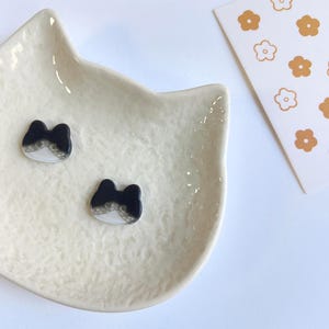 May include: Two small, black and white cat-shaped buttons with a smooth finish, resting inside a cream-colored, cat-shaped ceramic dish. Beside the dish is a white card featuring a repeating pattern of small, orange and white flower icons.