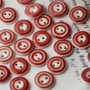 May include: A collection of red and white ceramic buttons, each with two holes. The buttons have a white centre with a red ring around it, and another red ring around the outer edge.