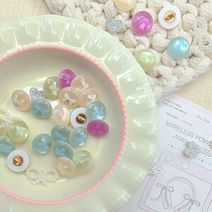 May include: An assortment of pastel beads and embellishments in shades of pink, blue, and iridescent colours, presented on a pale yellow dish with a pink rim. A small paper with the text "WIRELESS POWER" is also visible.