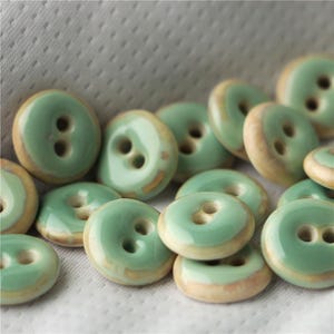 May include: A collection of green ceramic buttons with a light brown rim. The buttons have two holes each.