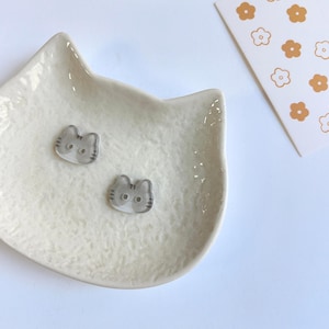 May include: Two small, translucent gray cat-shaped buttons with dark gray striped details and two holes in the center, resting inside a textured, off-white ceramic dish shaped like a cat head, next to a white card with orange flower icons.
