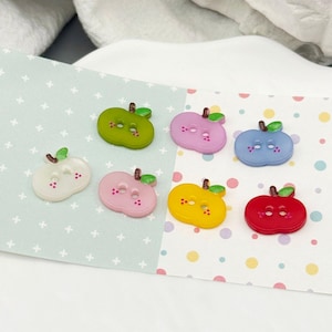 Può includere: Una collezione di sette bottoni a forma di mela in vari colori, tra cui verde, rosa, giallo, rosso, blu e bianco. Ogni bottone ha una piccola foglia verde e due asole. I bottoni sono disposti su una superficie a fantasia.