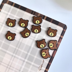 May include: A collection of ten small, brown bear-shaped buttons. Each button features a dark brown head with a lighter tan muzzle, two small black eyes, and a black nose and mouth. The buttons have two holes for attachment, positioned centrally.