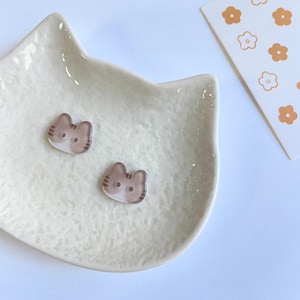 May include: Two small, flat, cat-shaped buttons featuring a brown and white tabby pattern with two holes for sewing. The buttons are resting inside a textured, off-white, cat-shaped ceramic dish next to a card with orange flower icons.