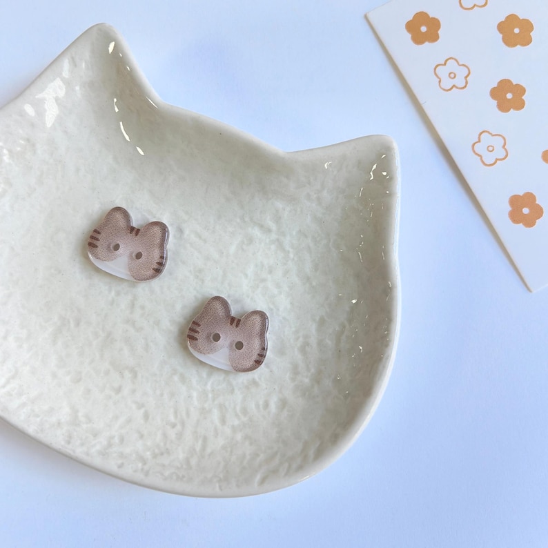 May include: Two small, flat, cat-shaped buttons featuring a brown and white tabby pattern with two holes for sewing. The buttons are resting inside a textured, off-white, cat-shaped ceramic dish next to a card with orange flower icons.