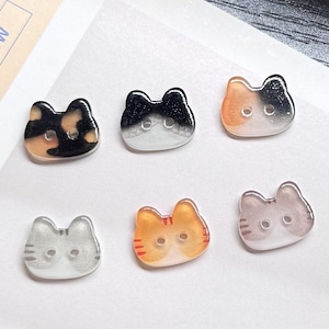 May include: A collection of six cat-shaped buttons in various colors and patterns. The buttons feature two holes for sewing and are made of a translucent material. Colors include black, white, orange, and gray, with some having striped or mottled designs.