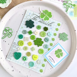 May include: A selection of green and white clover-themed embellishments, including buttons, stickers, and paper cutouts, arranged on a white tray. The text "The miracle" is visible on a patterned background. A card with the text "Clover Morning" is also present.