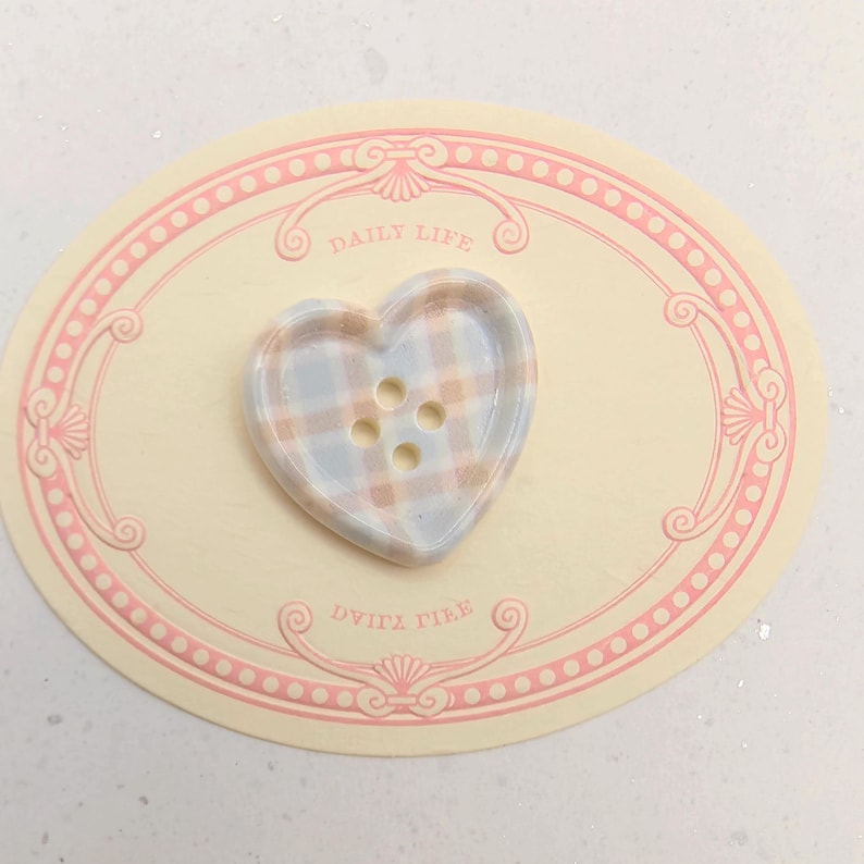 Op de afbeelding: Een hartvormige knoop met een ruitpatroon in blauw, beige en wit. De knoop heeft vier gaatjes en ligt op een cr&egrave;mekleurige ovale kaart met roze rand en de woorden "DAILY LIFE" en "DVITZ TIEE".