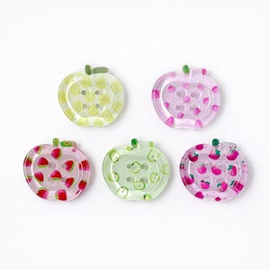 Può includere: Cinque bottoni a forma di mela in vari colori. I bottoni presentano disegni di frutta, tra cui limoni, fragole e mele. Ogni bottone ha un gambo verde e piccoli fori per cucire. I bottoni sono in plastica trasparente.