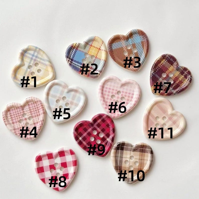 Op de afbeelding: Een verzameling van elf hartvormige knopen met verschillende ruitpatronen en kleuren, waaronder geel, blauw, roze en rood. Elke knoop heeft een uniek ontwerp en is genummerd van #1 tot #11. Perfect voor knutselprojecten.