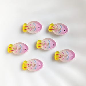May include: Six fish-shaped buttons. The buttons have a pink body, yellow tail, and blue accents. Each button has two holes for sewing. The buttons are arranged on a white surface.
