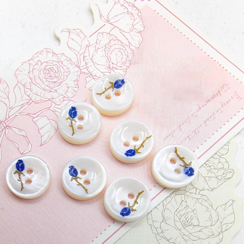May include: Seven white, round buttons with a pearlescent sheen. Each button features a blue floral design with gold accents. The buttons have two or three holes for sewing. The background is a pink card with floral illustrations.