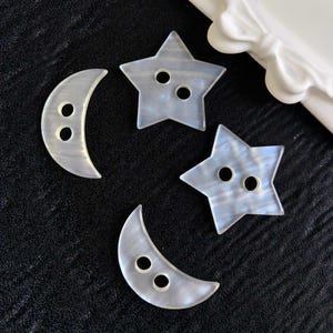 May include: Four iridescent buttons shaped as moons and stars. Each button has two holes for sewing. The buttons are pearlescent white with a subtle blue and grey shimmer. The buttons are on a black surface.