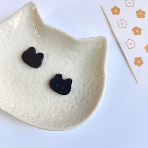 May include: Two small black buttons shaped like cat heads with two holes each, resting inside a cream-colored ceramic dish shaped like a cat head. A white rectangular card with a pattern of small orange and white flower outlines is visible.