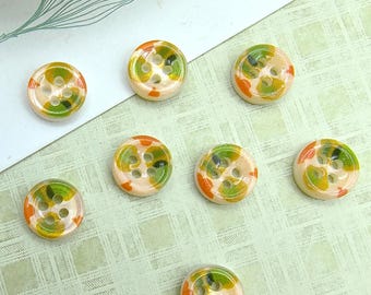 Può includere: Sette bottoni rotondi a quattro fori con una base traslucida color bianco sporco, caratterizzati da macchie irregolari e screziate di verde vibrante e arancione brillante. Ogni bottone presenta una superficie liscia, lucida e circolare.