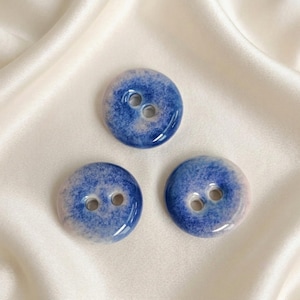 May include: Three circular buttons featuring a glossy finish with a gradient pattern transitioning from a deep blue center to a lighter, off-white edge. Each button has two centered holes for attachment and a smooth, reflective surface texture.