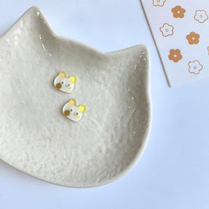 May include: Two small, translucent, cat-shaped buttons with yellow ears and a single brown dot on one cheek. These buttons are resting inside a white, textured, cat-shaped ceramic dish, positioned next to a white card with a brown flower pattern.