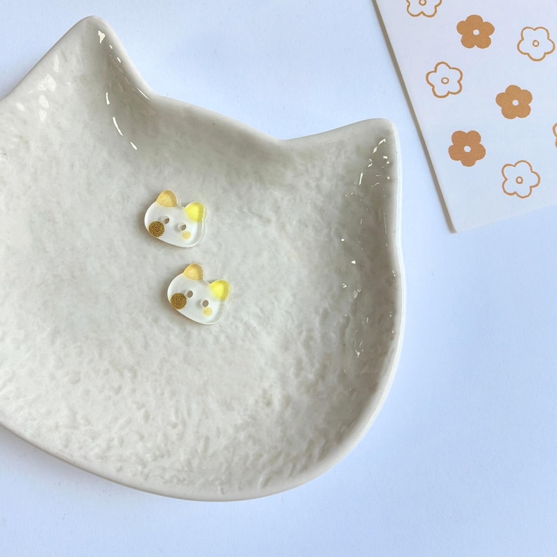 May include: Two small, translucent, cat-shaped buttons with yellow ears and a single brown dot on one cheek. These buttons are resting inside a white, textured, cat-shaped ceramic dish, positioned next to a white card with a brown flower pattern.