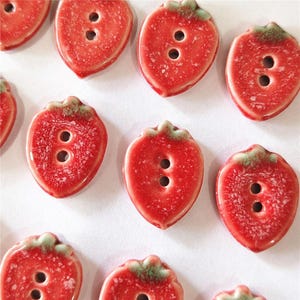 May include: A collection of red ceramic strawberry-shaped buttons with two holes each. The buttons have green tops and a glossy finish, perfect for sewing or crafting projects. The buttons are arranged on a white surface.
