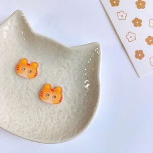 May include: Two small orange and yellow cat-shaped buttons with red striped details and two holes in the center. The buttons are placed on a white cat-shaped ceramic dish with a textured surface, next to a white card featuring a brown flower pattern.