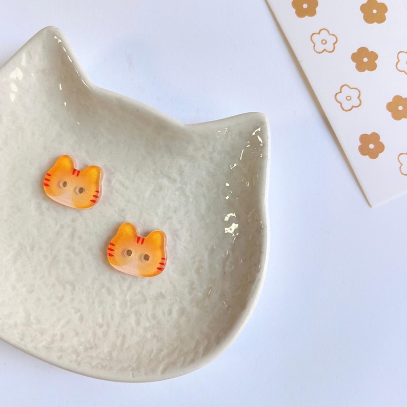 May include: Two small orange and yellow cat-shaped buttons with red striped details and two holes in the center. The buttons are placed on a white cat-shaped ceramic dish with a textured surface, next to a white card featuring a brown flower pattern.