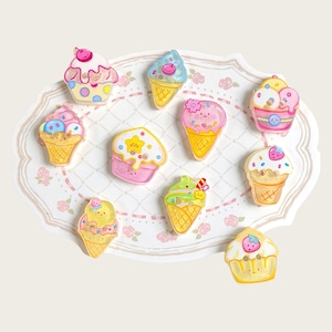 May include: A collection of ten small, flat, decorative buttons shaped like various ice cream cones, cupcakes, and desserts. Each button features pastel colors, including pink, yellow, blue, and green, with painted details like sprinkles, cherries, and smiling faces.